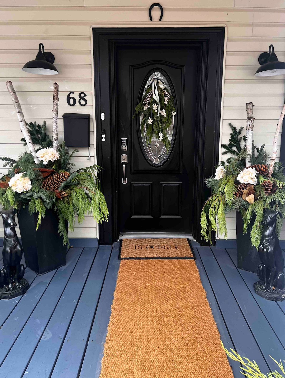 The entrance features a black door with an ornate oval glass window. Flanking the door are decorative planters filled with greenery and white floral arrangements. A coir doormat welcomes guests, and a jute runner leads up to the door, all complemented by stylish outdoor lighting.