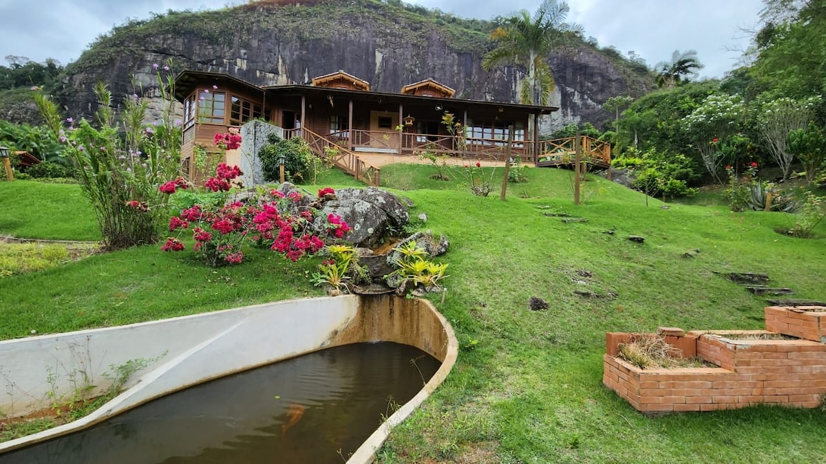 A charming wooden house is set against a backdrop of a rocky hill. Lush green grass surrounds the property, dotted with colorful flowers. A small water feature is visible in the foreground, enhancing the natural ambiance of the outdoor space.