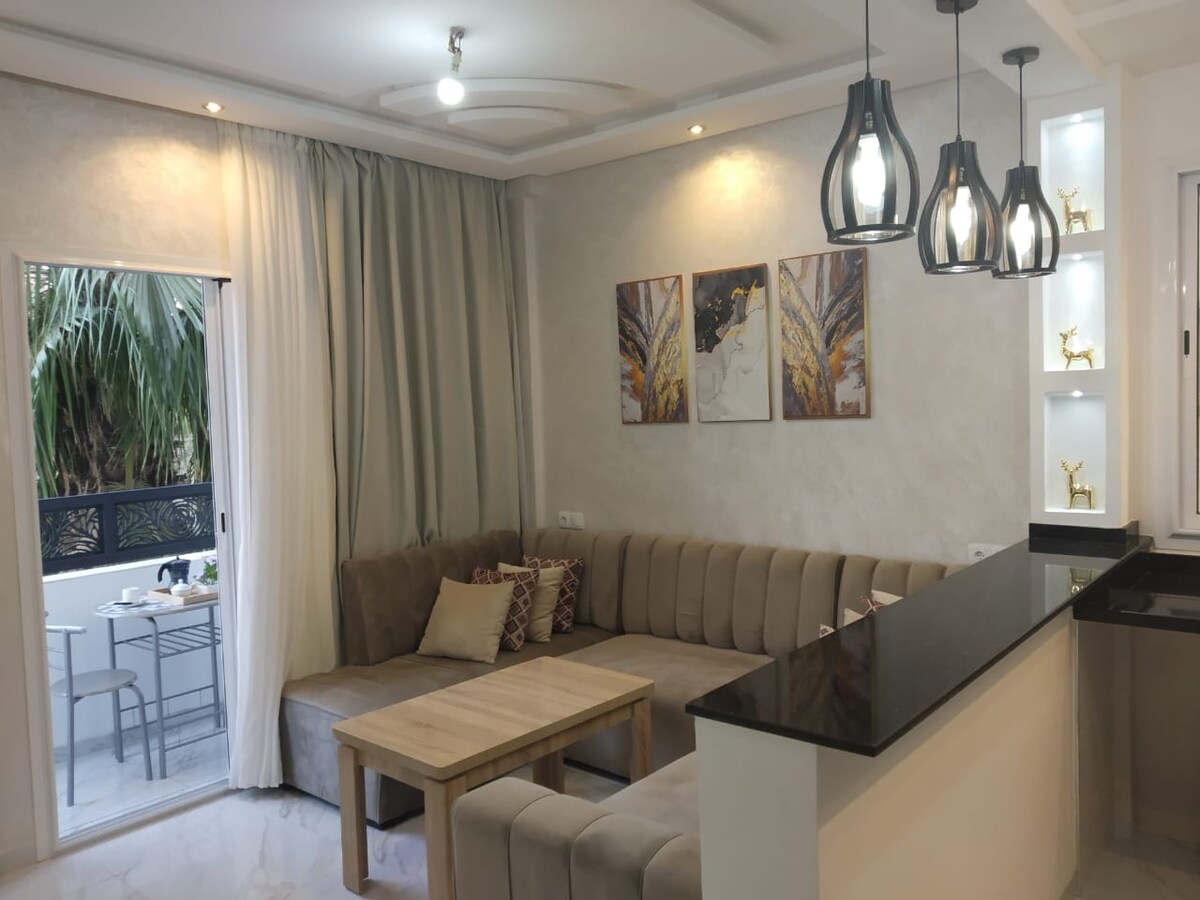 A spacious living area features a light-colored sectional sofa, complemented by a wooden coffee table. Modern light fixtures illuminate the room, which includes three pieces of wall art. A sliding glass door opens to a small outdoor seating area, framed by soft drapes.