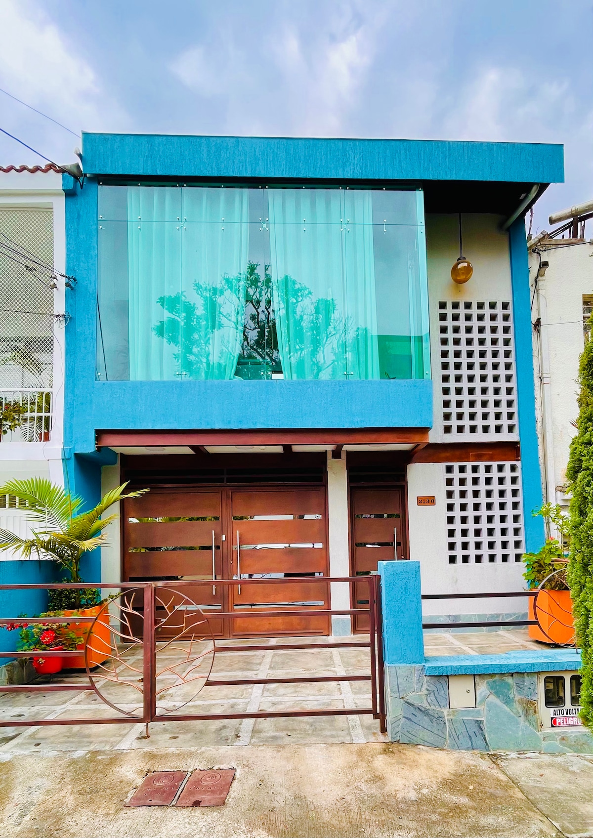 The exterior of the property showcases a modern design with a striking blue facade and large glass windows. The entrance features double wooden doors framed by decorative plants in terracotta pots. A clean walkway leads up to the door, emphasized by the bright surroundings.