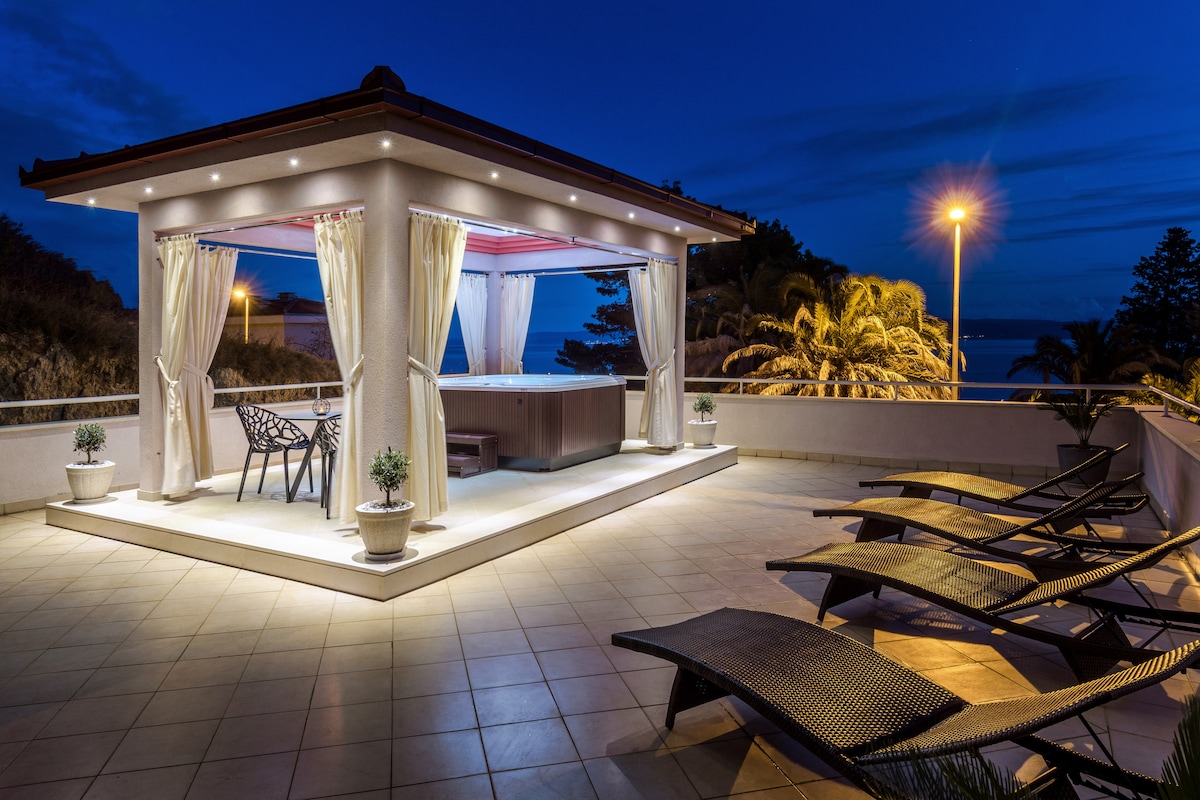 A spacious terrace is seen under a night sky, featuring a hot tub and soft seating area with draped curtains. Lounge chairs are positioned around the space, complemented by potted plants. Soft lighting enhances the inviting atmosphere, while glimpses of the sea can be observed in the background.