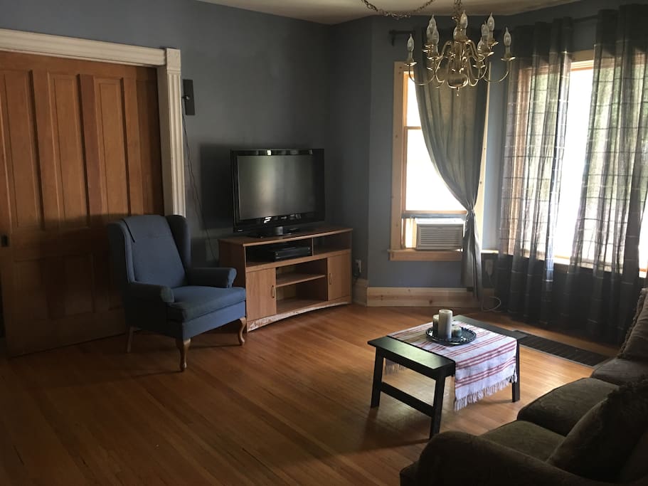 The Blue Room, the living room has hardwood floors (as does every room, except the tiled bathroom, and the carpeted Teal Room). There is a TV, window AC, dimmable chandelier, couch and easy chair, bay window, and buffet. The double sliding parlor door goes to the Goldenrod Bedroom.