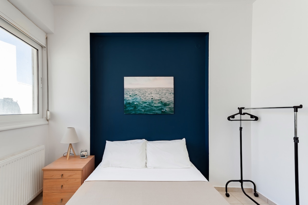 A simple bedroom is presented with a spacious bed covered in white linens. A blue accent wall features a serene ocean-themed artwork. Natural light enters through a window, while a wooden nightstand and a clothing rack complete the room's furnishings.