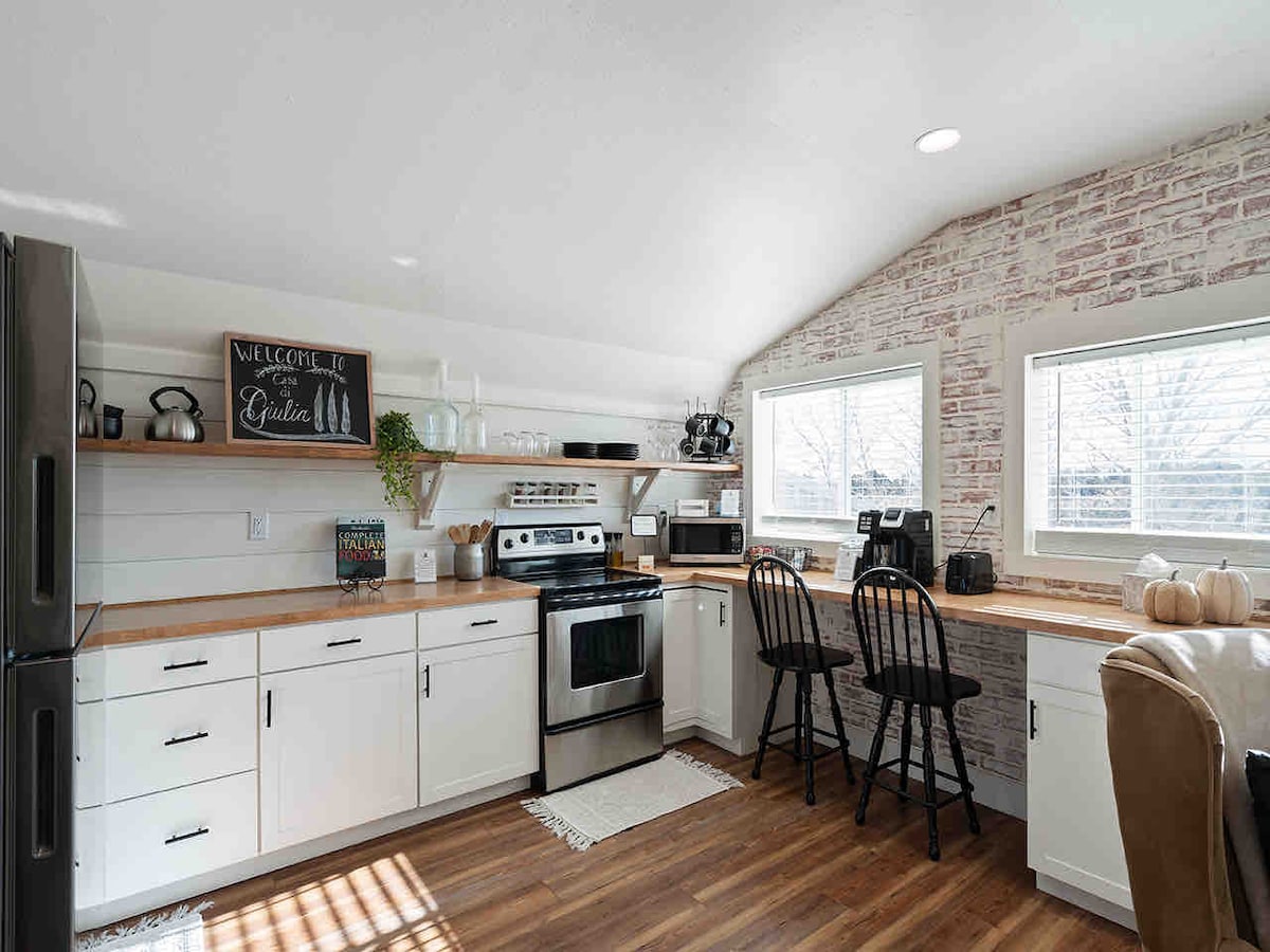 An airy kitchen is displayed with modern white cabinetry and wooden countertops. Stainless steel appliances are arranged alongside a coffee maker and additional kitchen equipment. Two black bar stools are situated at a raised counter, and natural light streams through the windows, enhancing the inviting atmosphere.