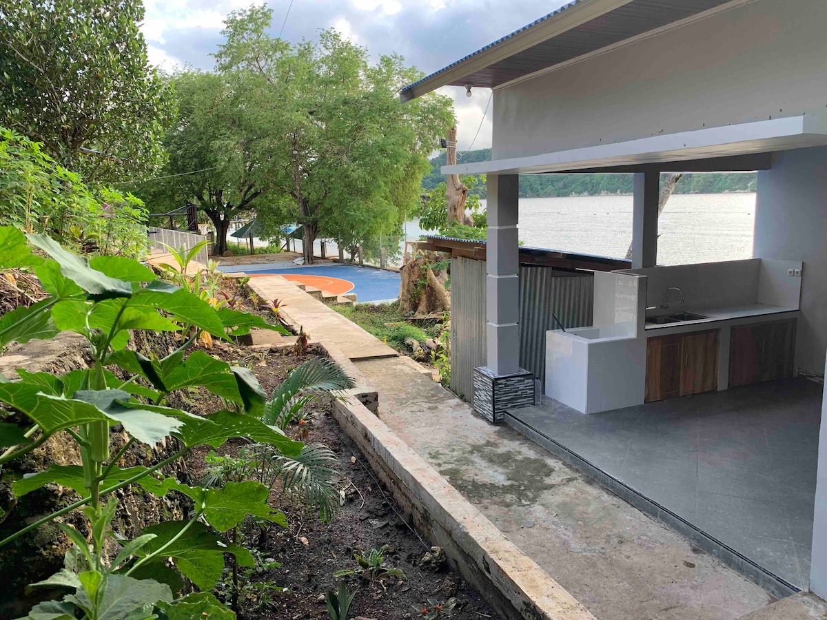A pathway leads from a landscaped area to a basketball half court near the water. An outdoor kitchen space features a simple design with a counter and seating. Lush greenery surrounds the area, enhancing the connection to nature.