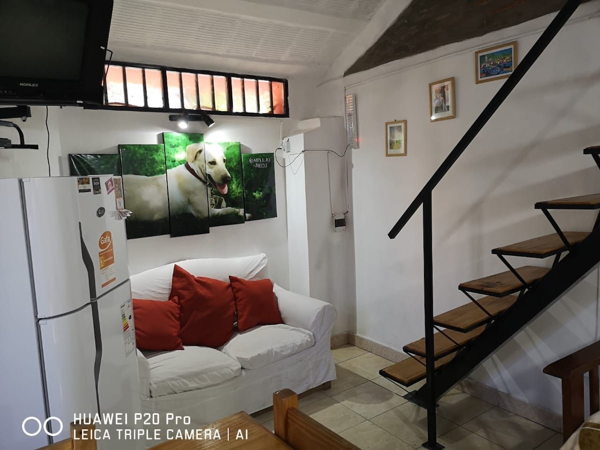 A cozy living area is equipped with a white couch adorned with red decorative pillows. A refrigerator stands in one corner, and a staircase leads to the upper level. On the wall, a large framed image of a dog adds character.