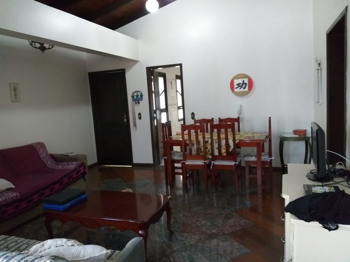 A spacious living area is highlighted by wooden flooring and natural light from overhead fixtures. A dark wooden coffee table sits in front of a comfortable sofa, while a dining table with colorful patterned chairs accommodates several guests. A television and decorative items are visible in the background.