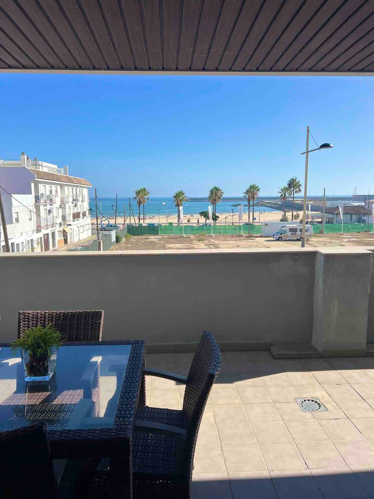 A terrace is shown featuring a dining table and woven chairs. In the background, a beach and palm trees are visible under clear blue skies. The view captures a serene coastal scene, with the ocean gently lapping against the shore.
