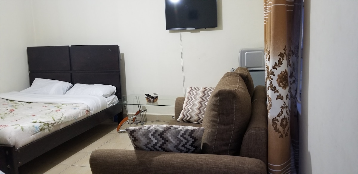 A cozy sleeping area features a spacious bed with floral bedding. A television is mounted on the wall above a small glass table. A brown sofa with patterned cushions is arranged nearby, contributing to the comfortable ambiance of the room.
