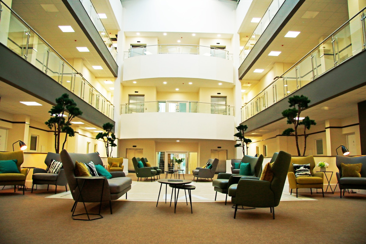 A spacious communal lounge area is featured, surrounded by multiple levels of seating arrangements. Green plants are placed strategically, and modern furniture in various colors enhances the contemporary design. Soft lighting complements the open space, creating an inviting atmosphere for relaxation.