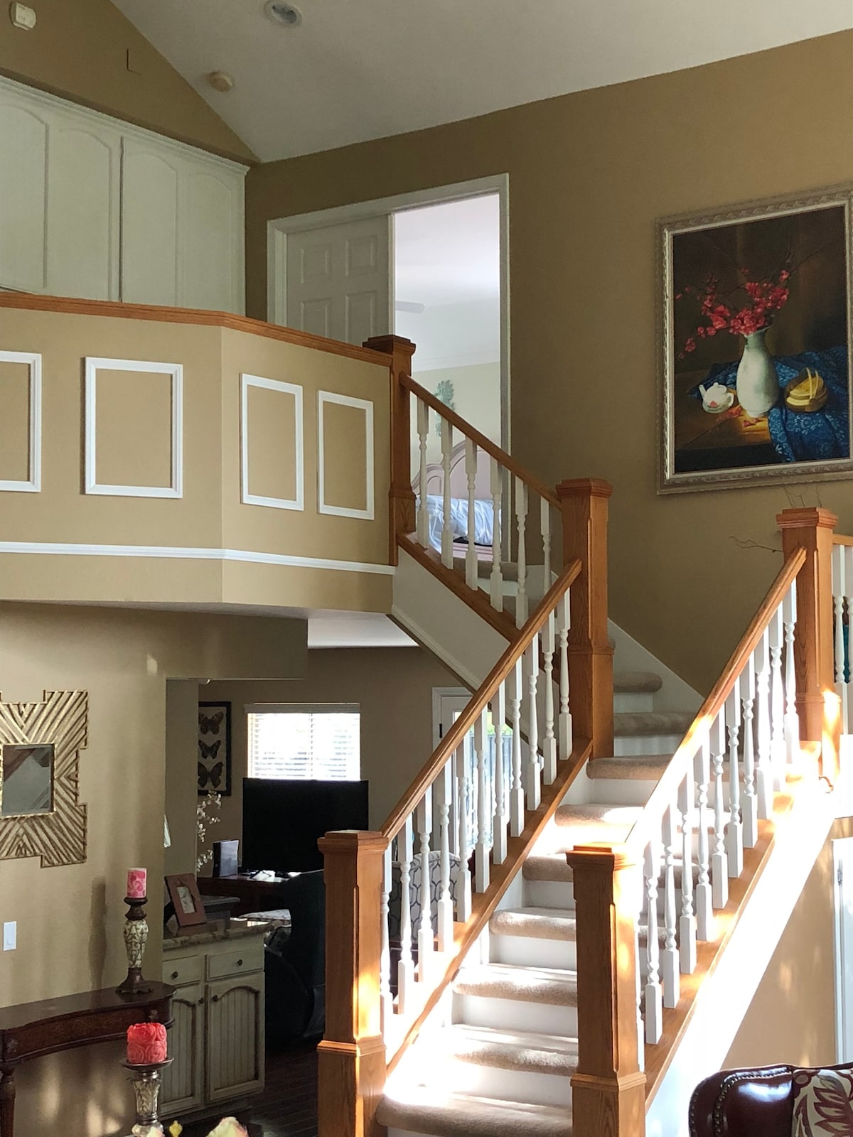 An elegant staircase with wooden railing leads to an upper-level landing. The walls are painted a warm beige, complemented by decorative features, including framed art. Soft lighting enters through a nearby window, enhancing the airy feeling of the space.