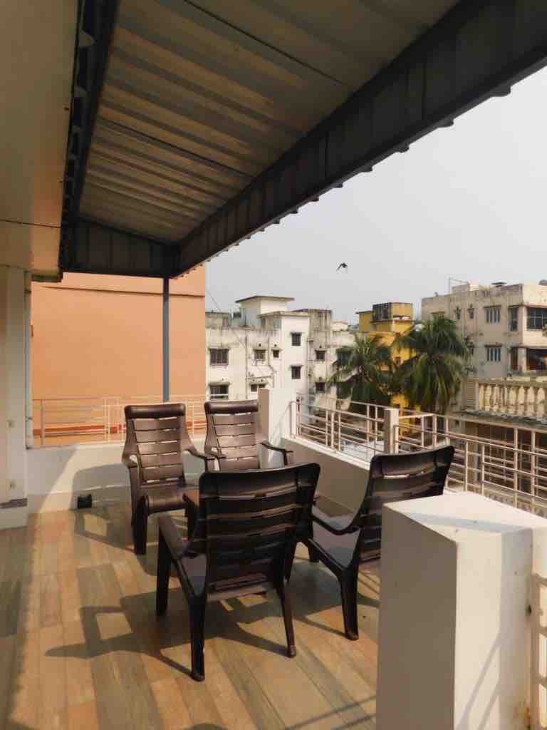 A balcony area is presented with a set of four black chairs arranged around a small table. The space features a tiled floor and is bordered by a railing, offering a view of nearby buildings and palm trees against a clear sky.
