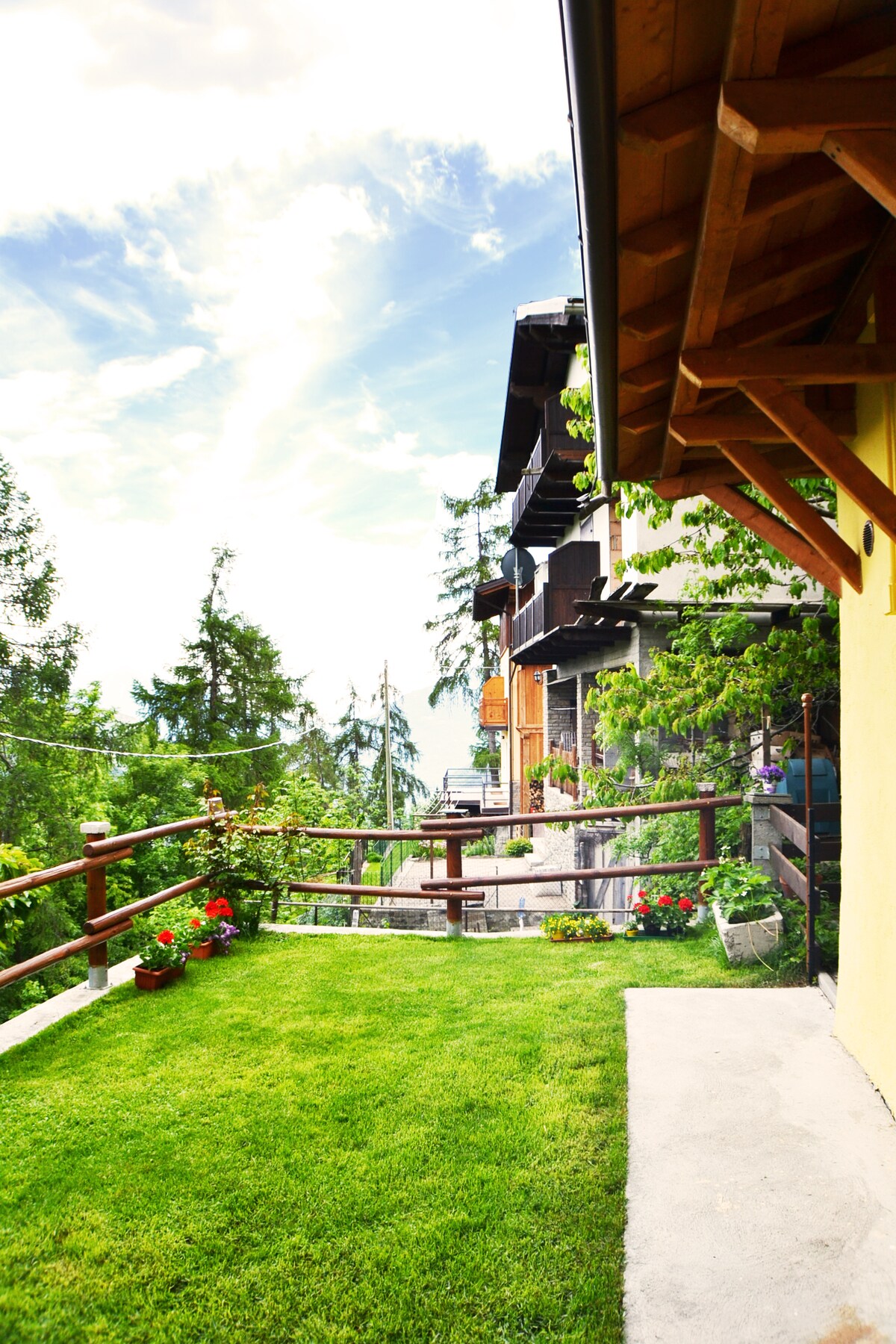 A well-maintained garden area is visible, featuring a neatly mowed lawn bordered by brightly colored flowers. Wooden railings surround the space, providing a view of the surrounding trees and mountains under a partly cloudy sky. The area is designed for relaxation outdoors.