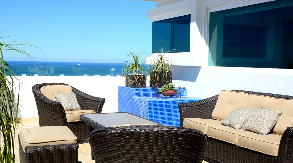 An outdoor seating area features comfortable rattan furniture with soft cushions. A table is centered amid the seating, while decorative plants add a touch of greenery. The expansive ocean view is visible through large windows, enhancing the tranquil ambiance of the terrace.