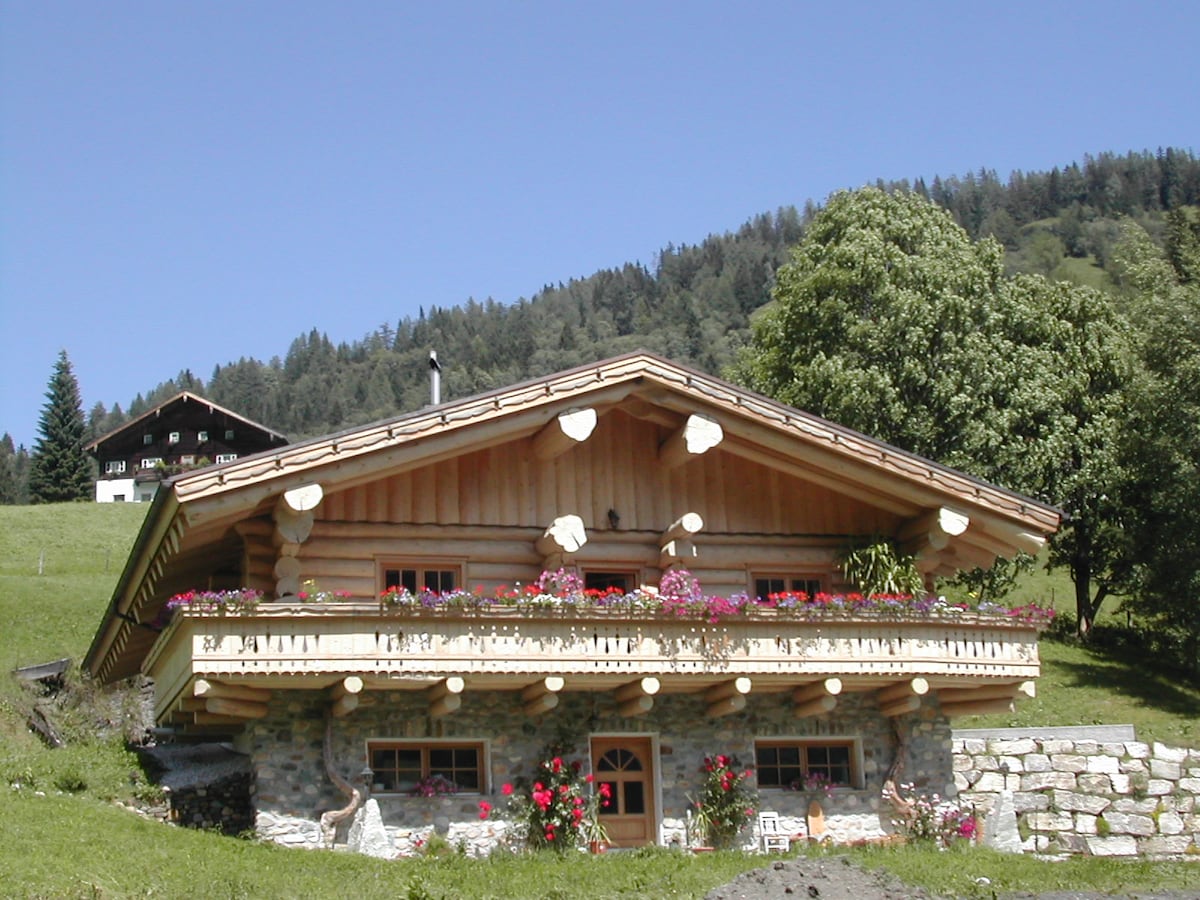 A charming blockhouse is set against a backdrop of green hills, featuring a wooden balcony adorned with vibrant flowers. The structure's rustic design includes a stone base and a sloped wooden roof, blending harmoniously into the surrounding natural landscape.