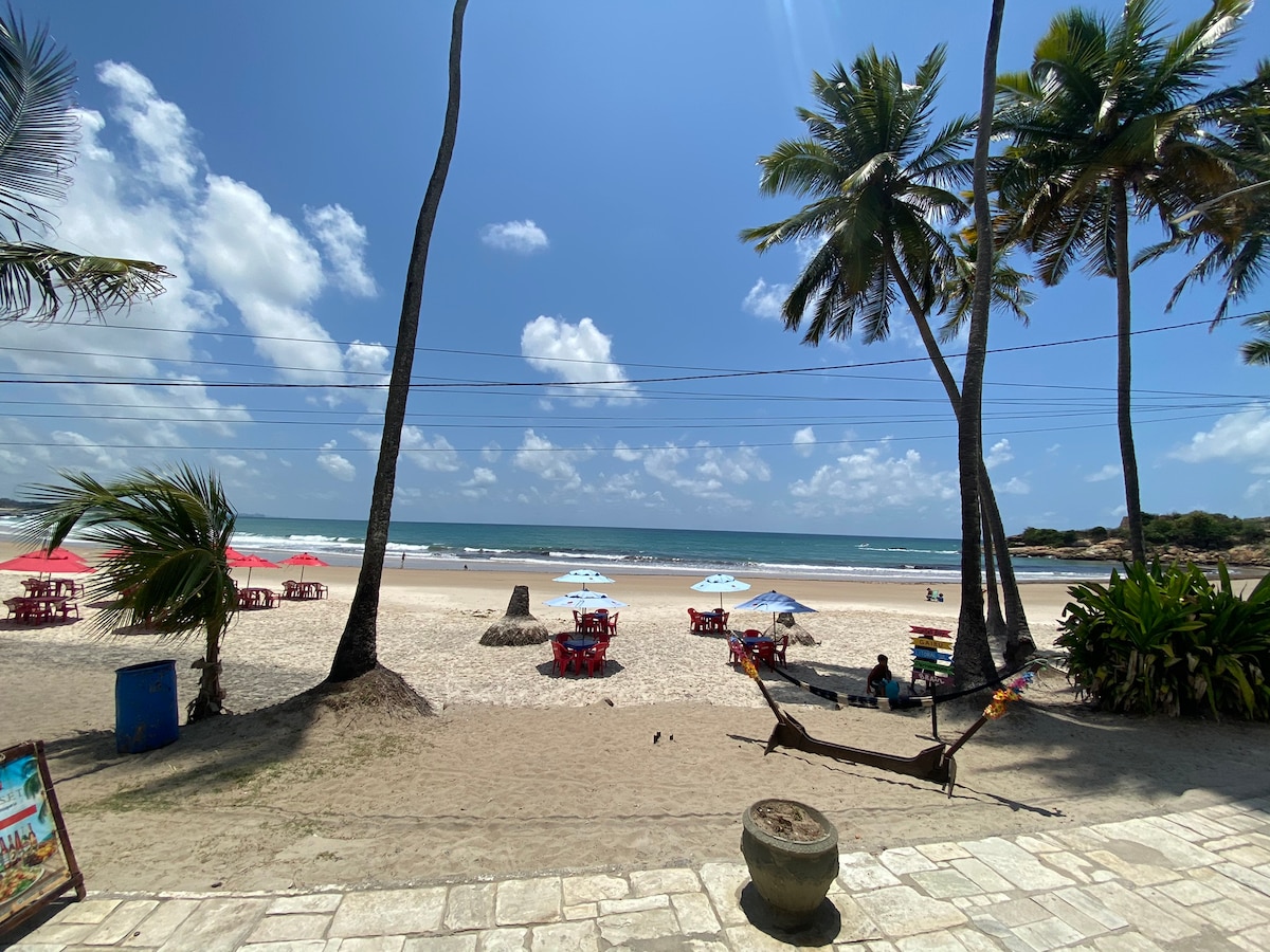 A view of the beach reveals a wide stretch of sand bordered by tall palm trees. Colorful umbrellas and chairs are arranged near the water, and gentle waves can be seen under a bright blue sky with scattered clouds.