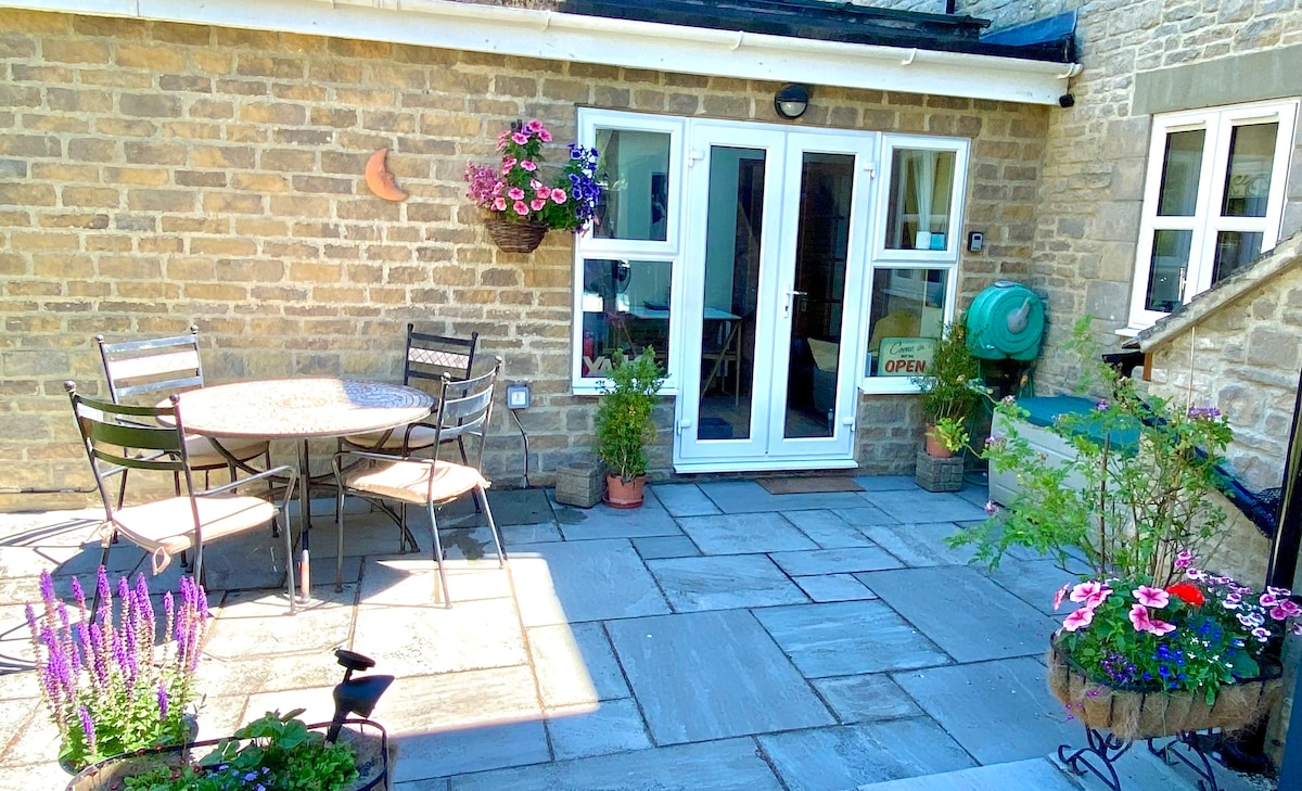 A private outdoor patio area features a round table surrounded by four chairs, complemented by potted plants and flowers. Sunlight reflects on the stone tiles, enhancing the inviting atmosphere. Glass double doors lead into the annex, providing easy access from the outdoor space.