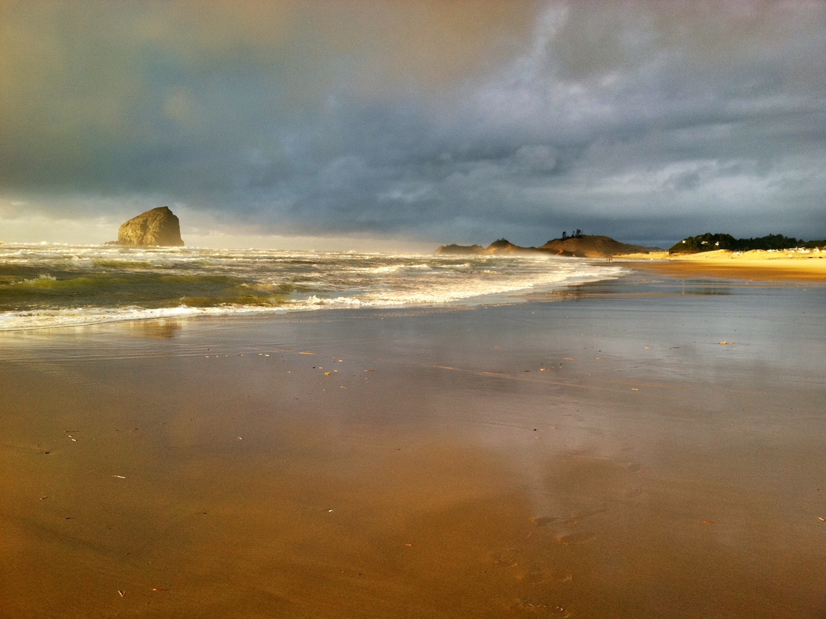 A serene beach scene captures the waves gently lapping the shore, with smooth wet sand reflecting the soft light. Haystack Rock is visible in the distance, framed by dark clouds and rolling hills along the coastline.