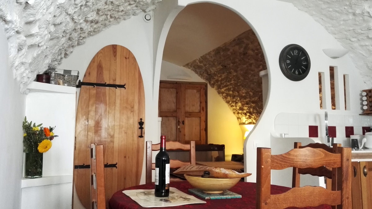 A cozy dining area features a rounded stone ceiling and an arched opening leading to another room. A wooden table is set with a bottle of wine and fresh flowers, while intricately designed doors and rustic furnishings add charm.