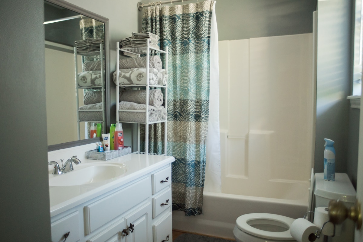 A clean and functional bathroom is equipped with a white vanity featuring a sink and storage drawers. A shower/tub combination is present alongside neatly arranged towels. A window allows natural light to illuminate the space, and a simple decorative curtain adds a touch of color.