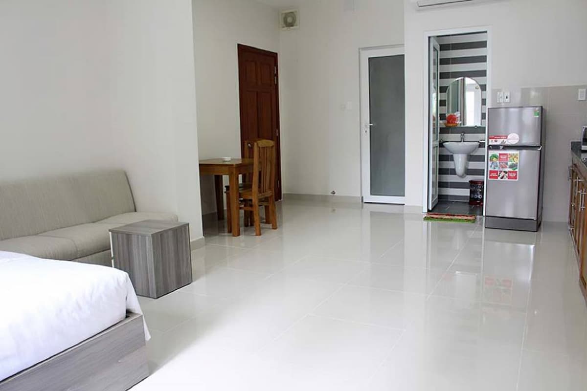 A studio unit is shown with a spacious layout featuring a light-toned sofa and a simple wooden dining table paired with chairs. A kitchenette and refrigerator are visible in the background, and the bathroom area is separated by a frosted glass door.