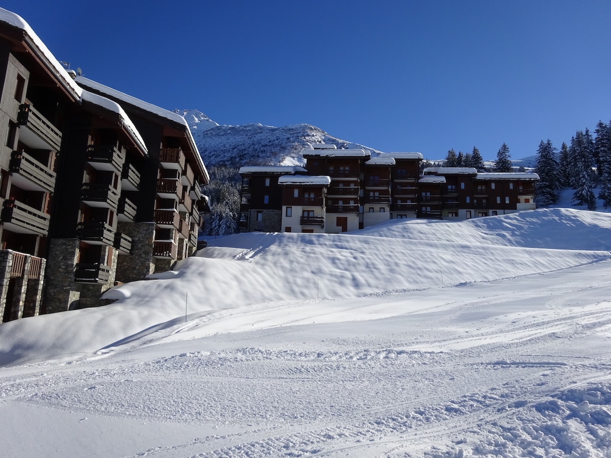 A snow-covered landscape is depicted, featuring multiple chalet-style buildings with wooden balconies. The clear blue sky contrasts with the white snow blanket that covers the ground, highlighting the peaceful alpine environment. In the background, snow-capped mountains add to the tranquil setting.