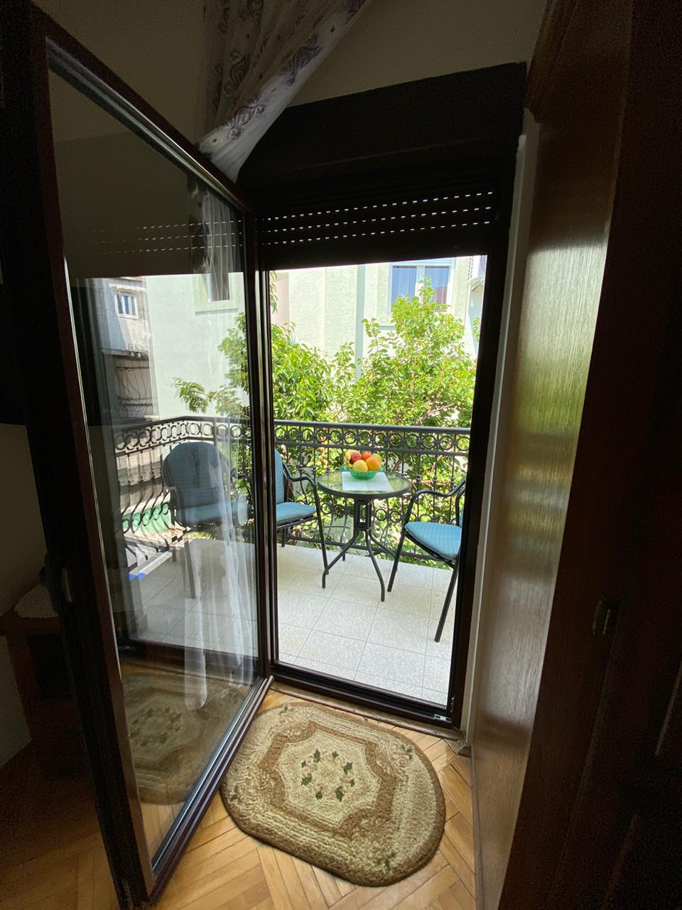 A set of glass doors opens to a small balcony featuring a round table and two chairs. The balcony is adorned with greenery, providing a serene view. A patterned rug lies on the floor inside, enhancing the transition from the interior to the outdoor space.