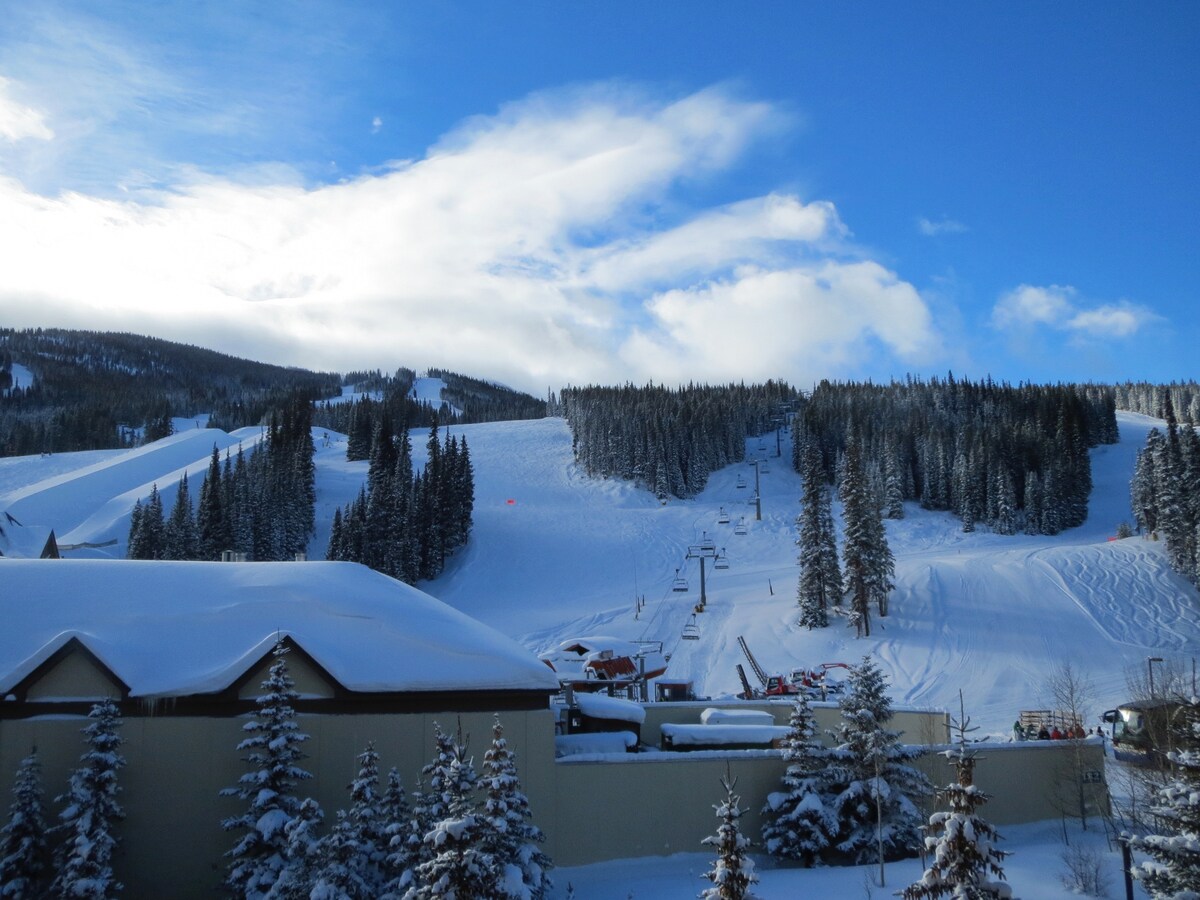A snowy landscape features well-groomed ski slopes and tall pine trees outlined against a clear blue sky. The scene captures the tranquil ambiance of the mountains, with vibrant white snow blanketing the ground and a glimpse of ski lifts in the distance.