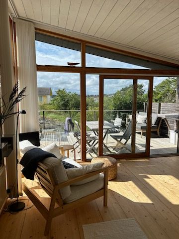 A light-filled living area features large glass doors opening to a balcony with outdoor seating. Natural wood tones are present in the furnishings and flooring. Lush greenery is visible through the windows, creating a serene connection to the surrounding landscape.