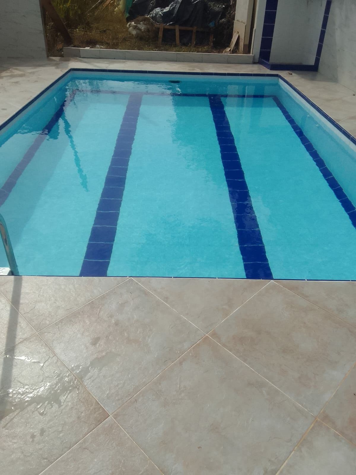 A clear swimming pool is visible, featuring a light blue surface marked by two darker blue lines. The pool is surrounded by tiled flooring, indicating a well-maintained outdoor area. Sunlight reflects off the water, enhancing the inviting appearance of the space.