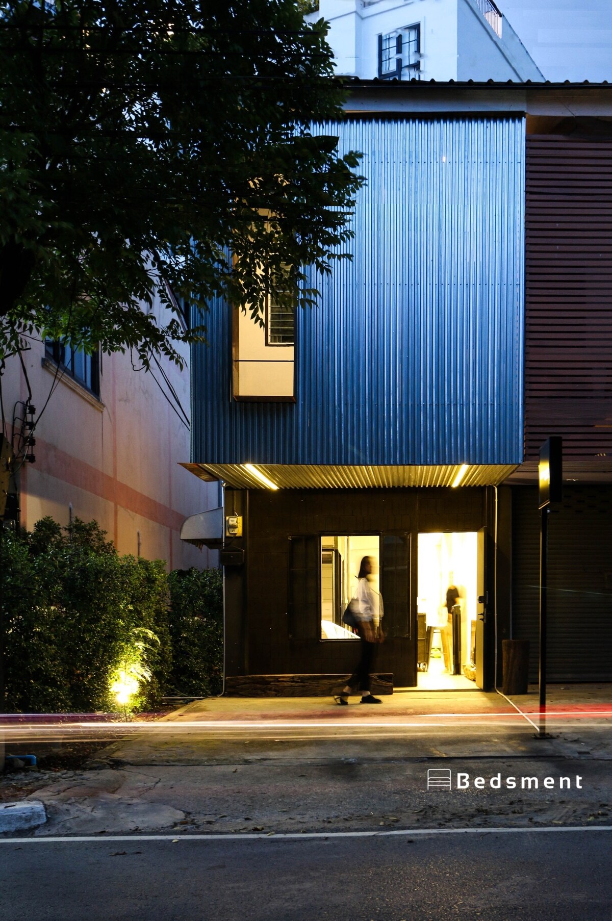 The modern two-story townhouse features a metallic facade and large glass entrance. Soft lighting illuminates the entryway, revealing guests entering the space. A glimpse of greenery is visible along the pathway, enhancing the welcoming ambiance of the property.