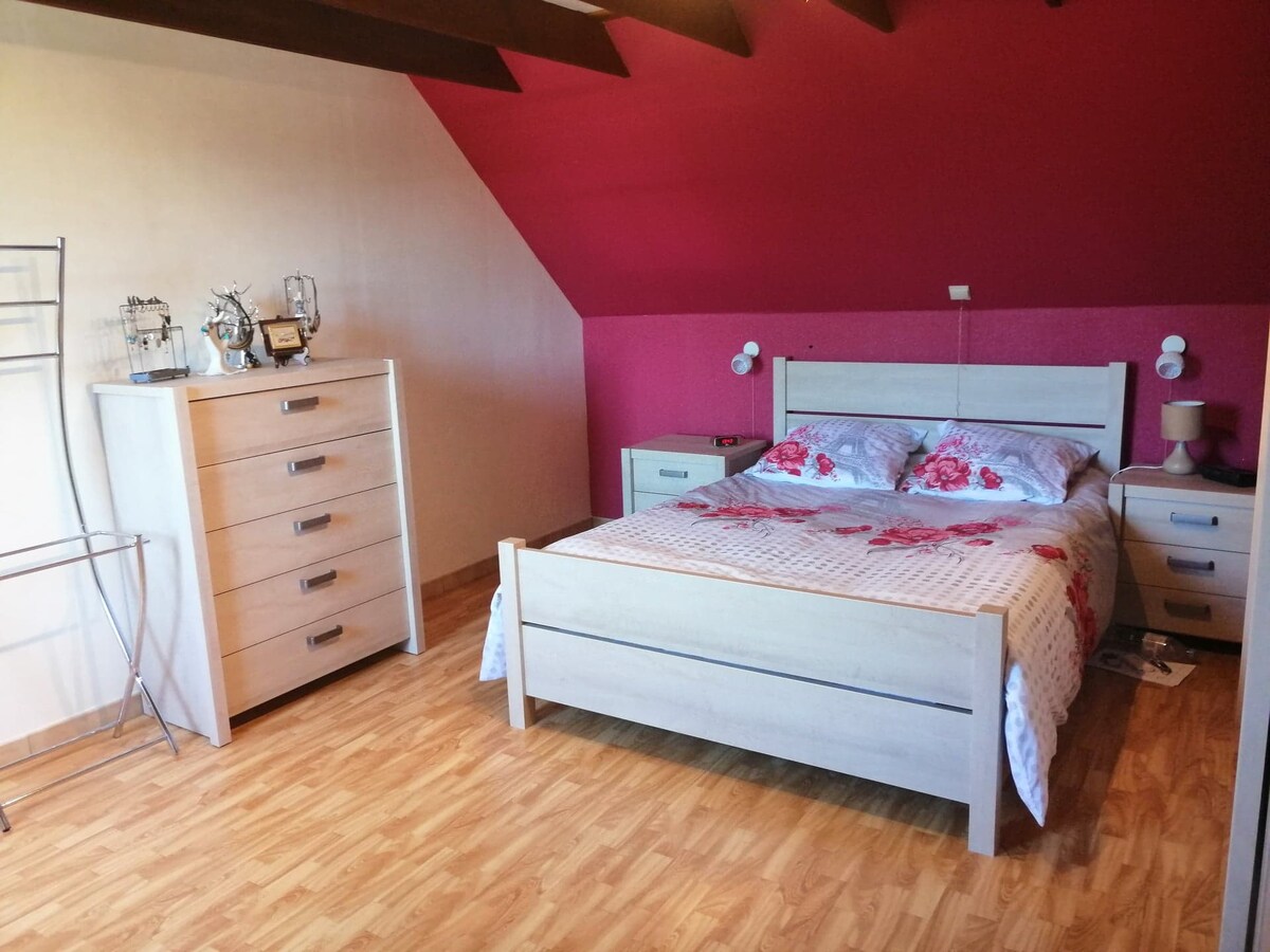 The room features a light wooden floor and slanted ceiling, with walls in soft pink and white. A spacious bed with floral bedding dominates the space, accompanied by a nightstand and a matching dresser. A clothing rack is also positioned against one wall.