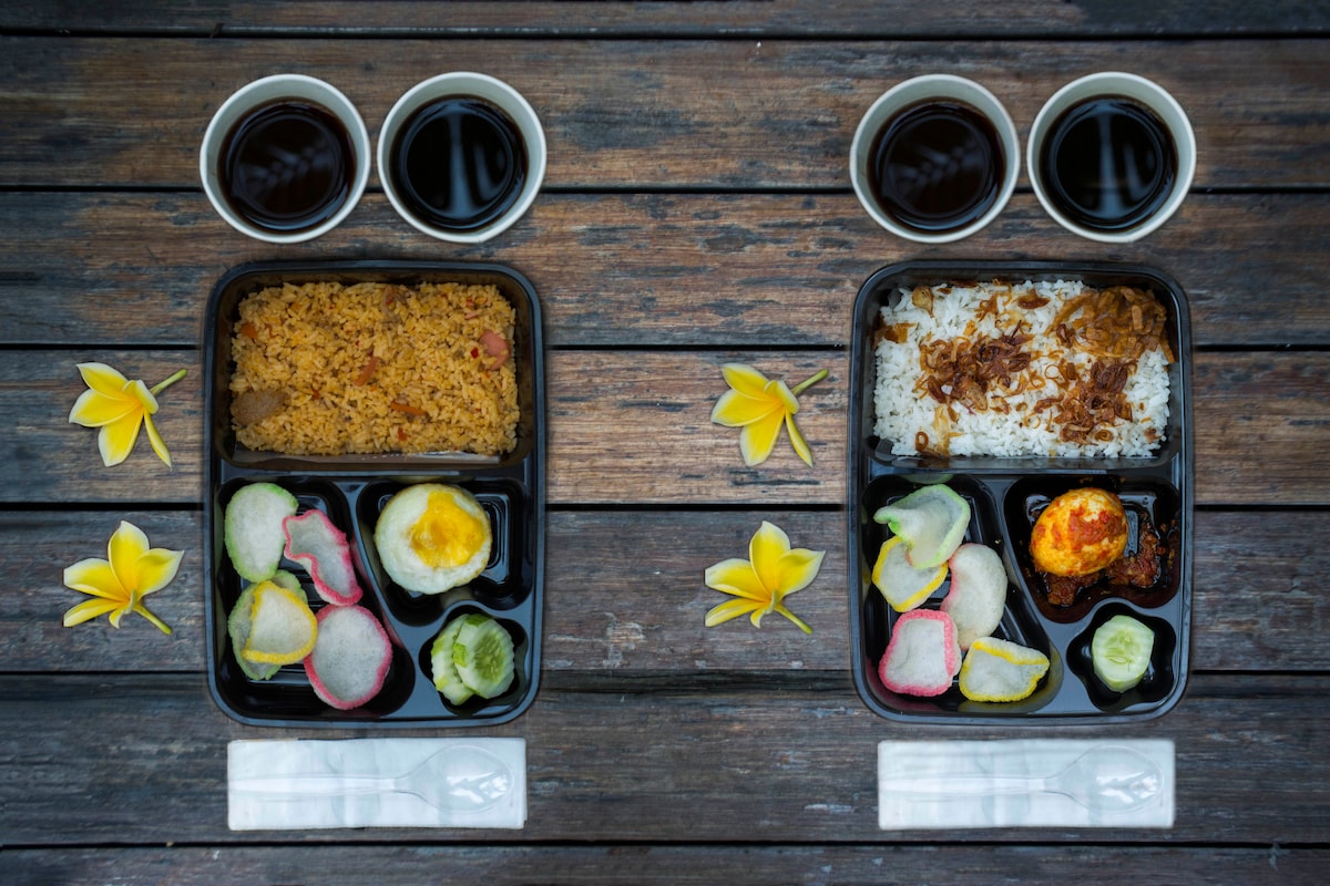 Two neatly arranged meal boxes are displayed on a wooden surface. Each box contains a serving of rice, vegetables, and a protein. Two cups of dark beverage sit nearby, complemented by yellow flowers placed around the meal, enhancing the dining experience.