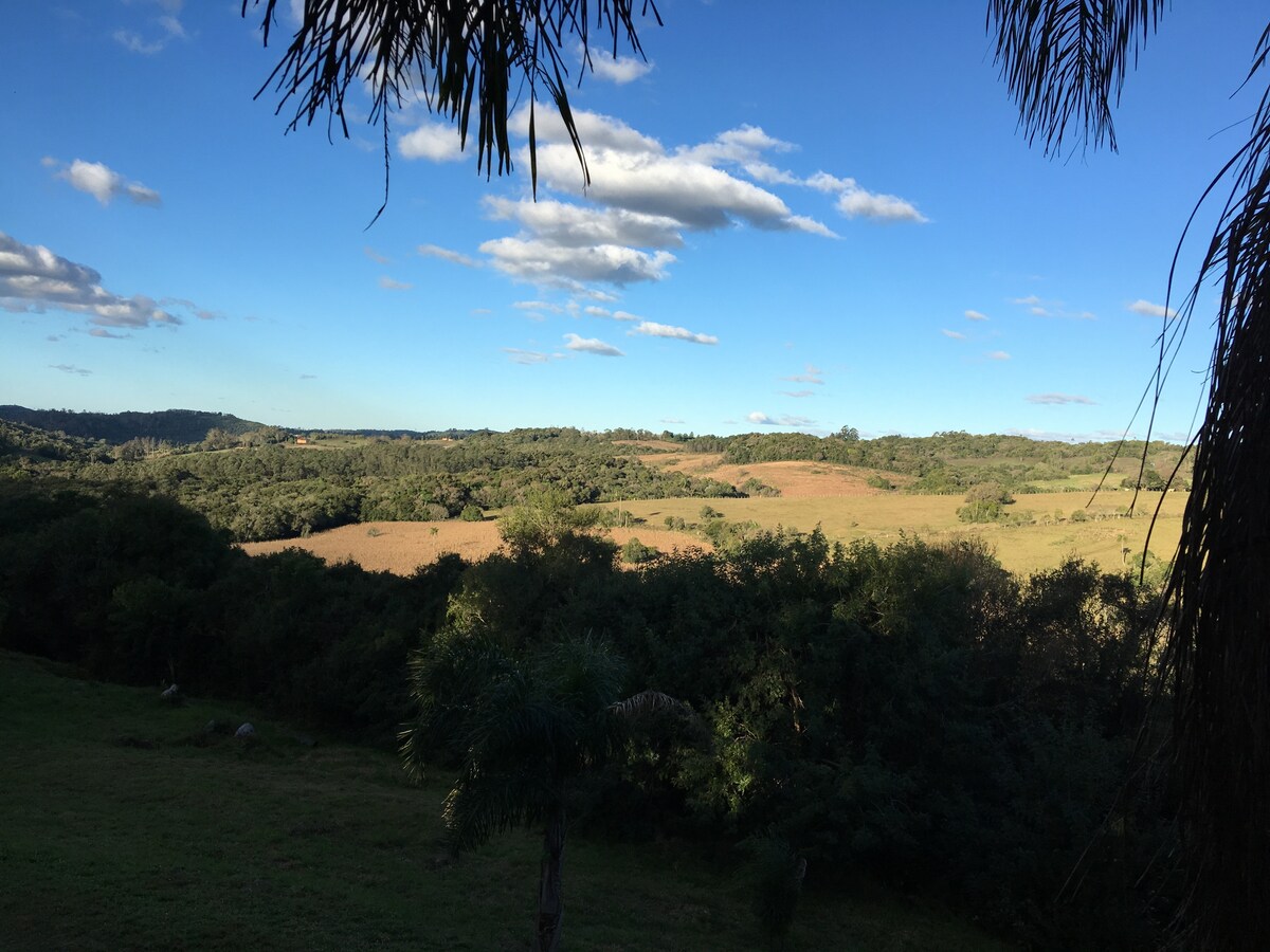 A panoramic view of rolling hills and lush greenery is captured under a clear blue sky with fluffy clouds. The landscape features a gentle mix of open fields and wooded areas, inviting tranquility and natural beauty.