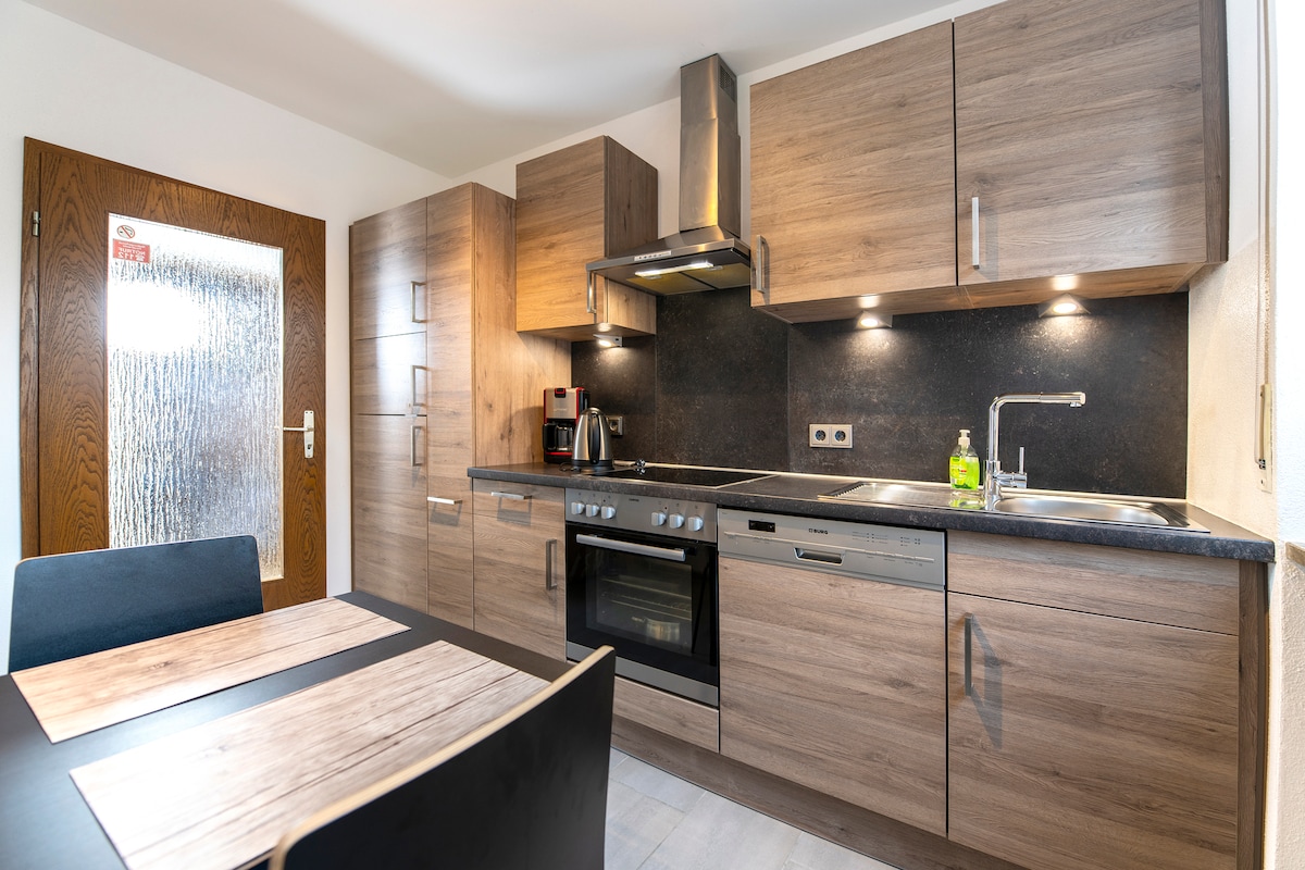 The kitchen features modern wooden cabinets with a sleek countertop. Appliances include an oven, stovetop, coffee maker, and dishwasher, all set against a dark backsplash. A dining table with dark chairs is positioned nearby, offering a cozy dining space.