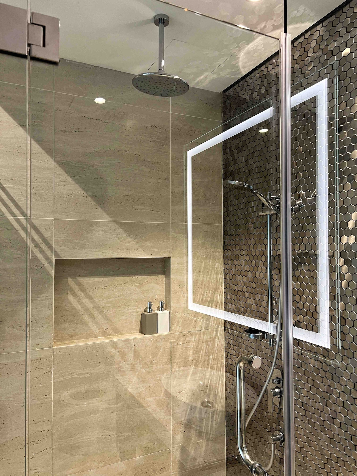 The bathroom features a modern glass shower enclosure with a rainfall showerhead. Elegant tiles cover the walls, highlighted by a backlit mirror. A recessed shelf holds toiletries, and the floor displays a continuous tile pattern, contributing to a streamlined aesthetic.