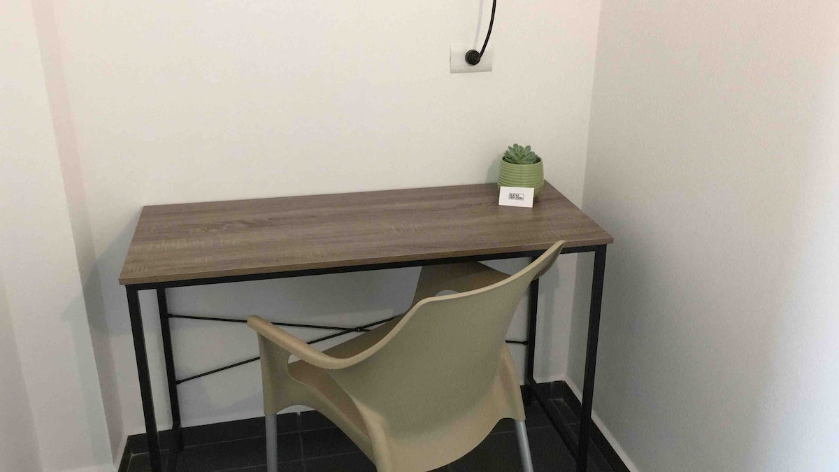 A minimalistic workspace is shown, featuring a wooden desk with a slender design and a modern chair. A small potted plant sits on the desk, alongside a nameplate. The walls are painted in a neutral tone, and the flooring is dark, providing contrast.