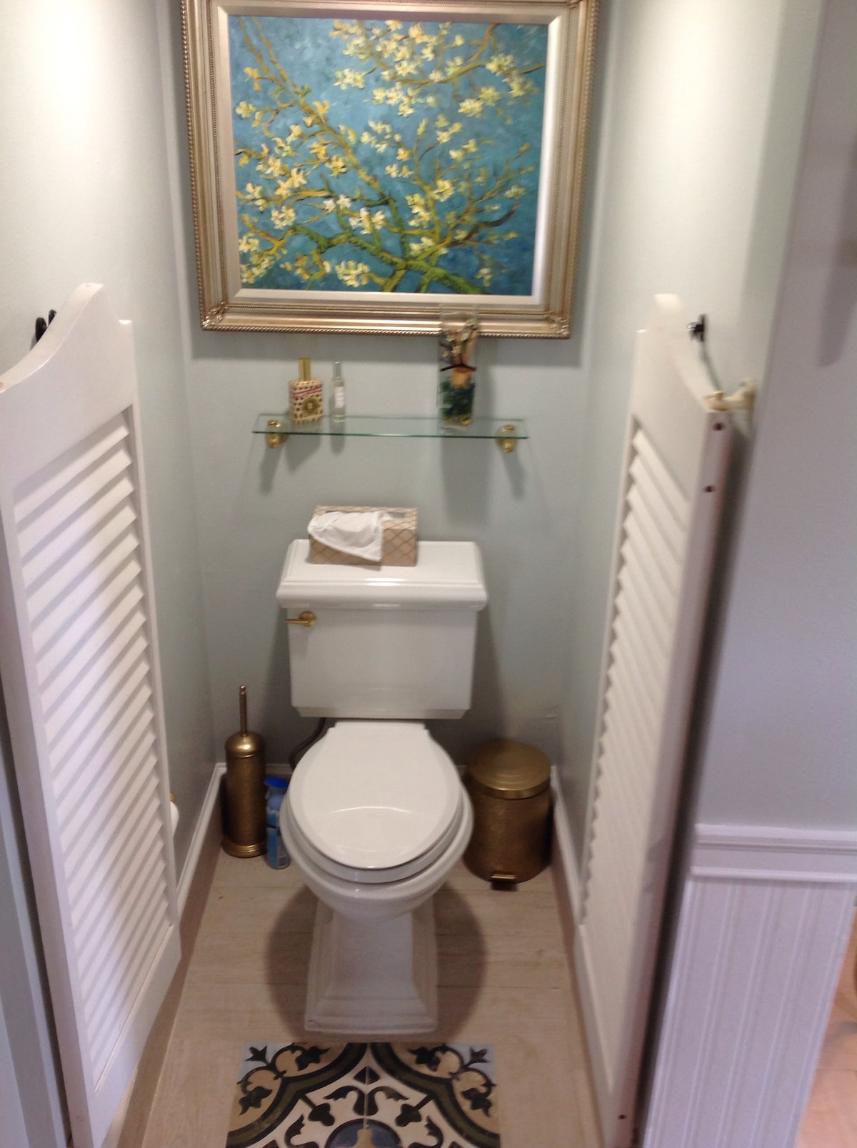 The bathroom features a compact layout with a toilet and a small decorative shelf above it. A piece of artwork with a floral theme hangs on the wall, complemented by a gold accent piece and toiletries arranged neatly. Light colors and elegant details enhance the space.