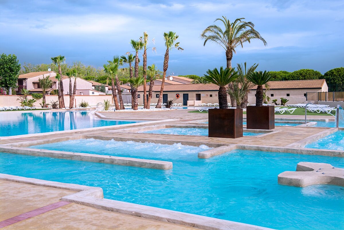A spacious pool area features various interconnected sections of water, surrounded by palm trees and lounge chairs. The tranquil blue water is enhanced by modern landscaping elements, creating an inviting outdoor space perfect for relaxation.