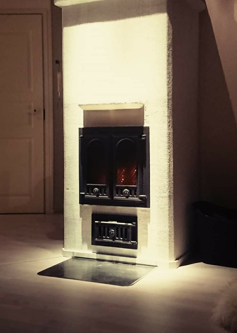 A classic stove is positioned against a textured wall, offering warmth with glowing embers visible through its arched panels. A metallic base reflects light, adding a subtle contrast to the surrounding space. A door is visible in the background, enhancing the room's overall layout.