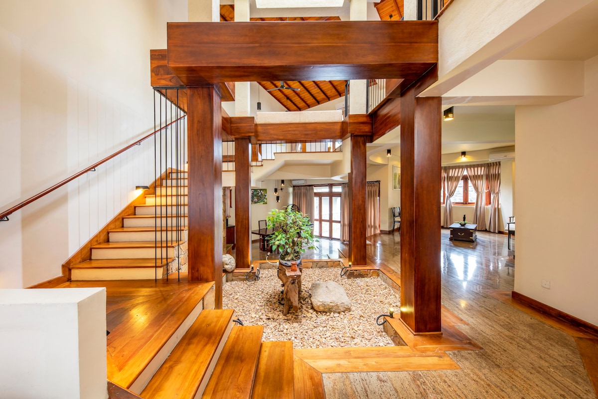 An open foyer features a central stone decor with a small tree and rocks, surrounded by polished wooden beams. Off to the side, a staircase leads to the upper level, while expansive windows illuminate the space with natural light.