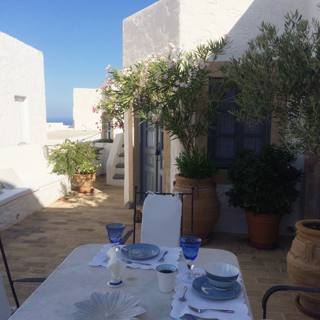 An outdoor dining area is set on a stone patio, featuring a table adorned with plates and glassware. Potted plants and flowers provide greenery, while whitewashed walls reflect the sun's light. A stairway leads to additional upper levels, enhancing the open and airy feel.