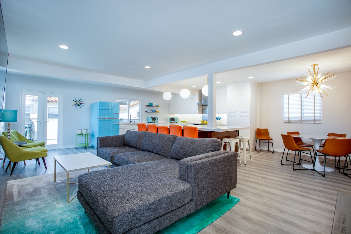An open-concept living area features a spacious sectional sofa in gray, complemented by vibrant orange and green chairs. A large dining table is visible, surrounded by stylish chairs. Modern kitchen appliances are highlighted, with natural light coming from windows and doors that lead to the outdoor space.