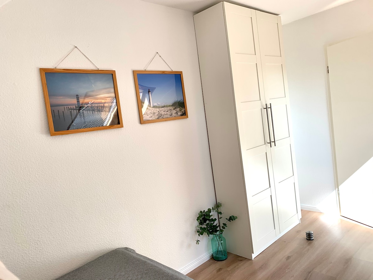 A bright corner of the room is highlighted by two framed photographs depicting scenic coastal views. A sleek, light-colored wardrobe stands against the wall, while a small green plant adds a touch of nature to the space.