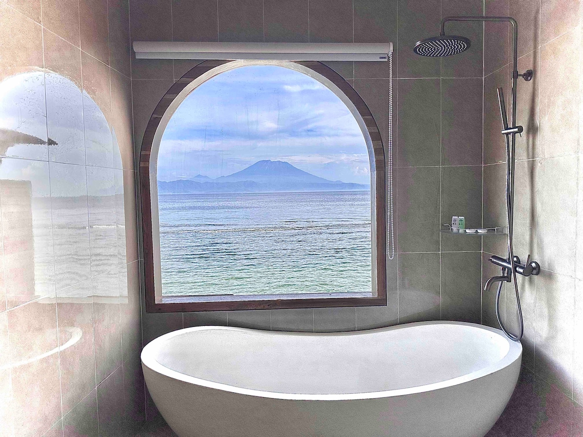 A freestanding bathtub is positioned in front of a large arched window, offering views of the ocean and distant mountains. Natural light fills the space, highlighting the light-colored tiles and modern fixtures. A showerhead is mounted above for convenience.