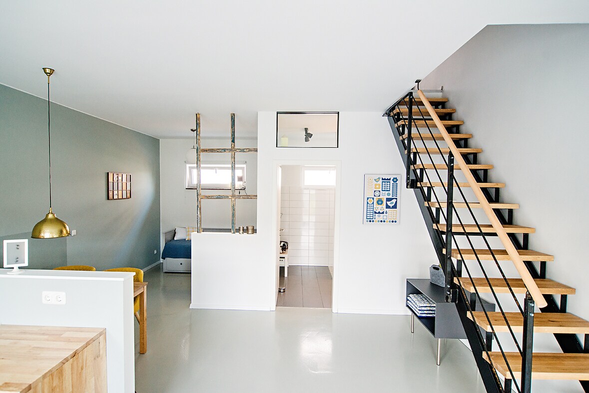 The spacious open layout features a sleek staircase with wooden steps, leading to the upper level. Natural light floods the room, highlighting the pale blue and gray walls. A dining area is visible alongside a cozy seating area, complemented by modern decor.