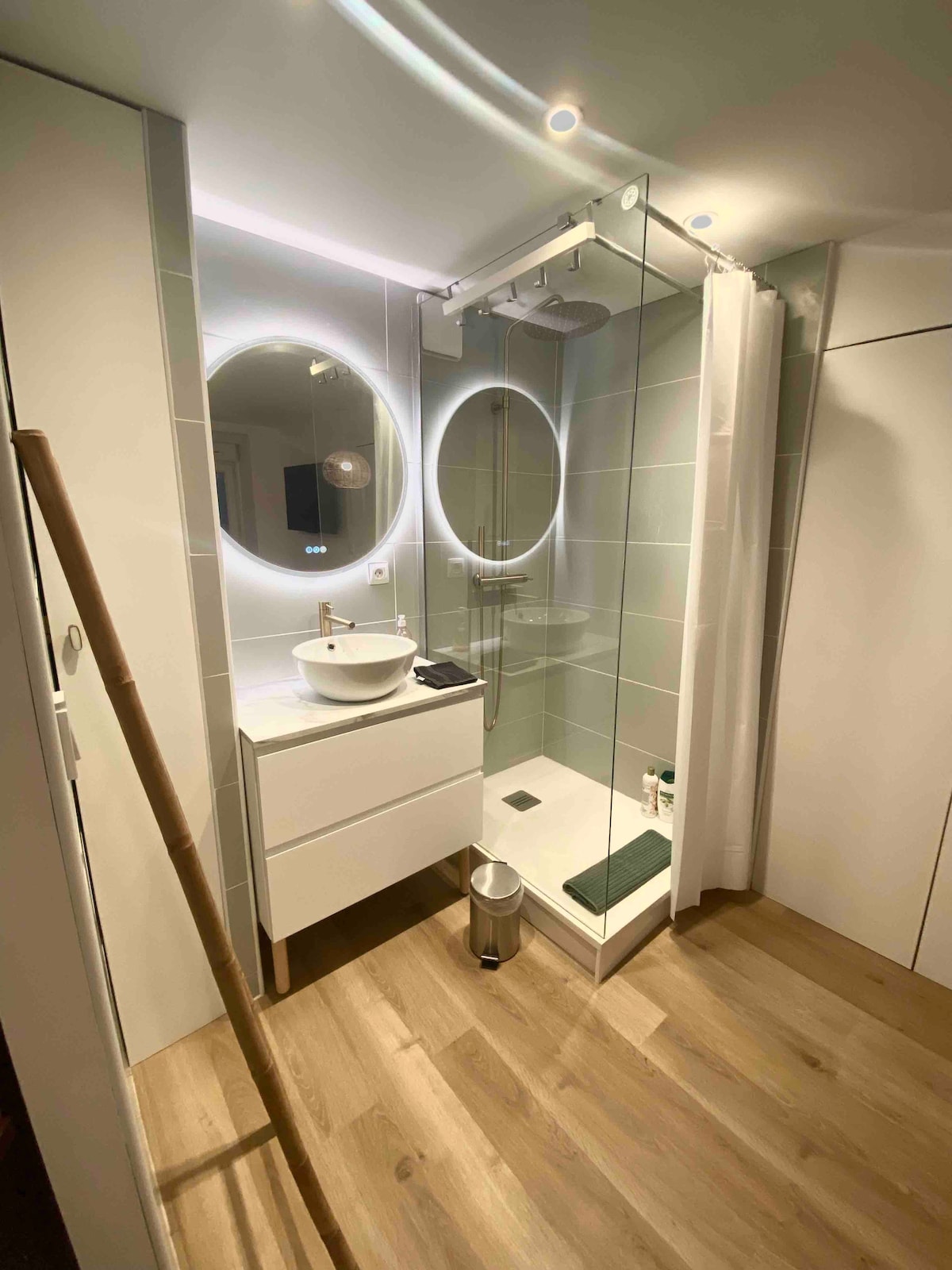 A modern bathroom is displayed, featuring a walk-in shower enclosed by glass panels. A circular mirror above a stylish sink reflects soft lighting. Light wooden flooring complements the clean, minimal design, while a small mat is positioned near the shower area.