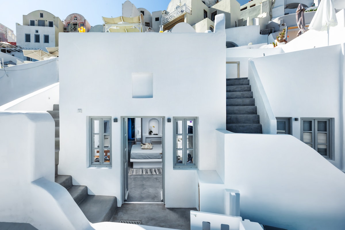 A whitewashed building is depicted, characterized by multiple levels and a minimalist design. Large windows framed in blue allow natural light to fill the interior spaces, while stairs lead to various entries and terraces. The surrounding architecture reflects traditional Cycladic style.
