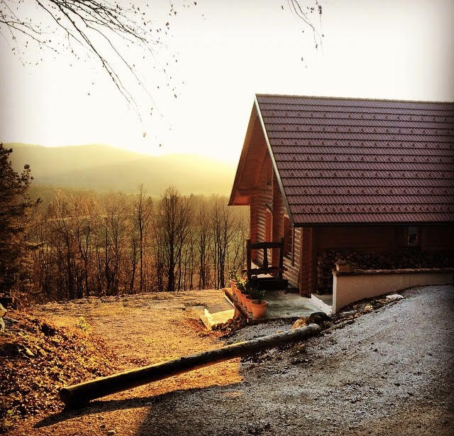 A rustic cabin features a sloped roof and wooden exterior nestled against a backdrop of rolling hills. Soft golden light radiates during sunset, casting gentle shadows on the gravel pathway leading to the cabin. The scene is framed by bare trees, creating a serene atmosphere.