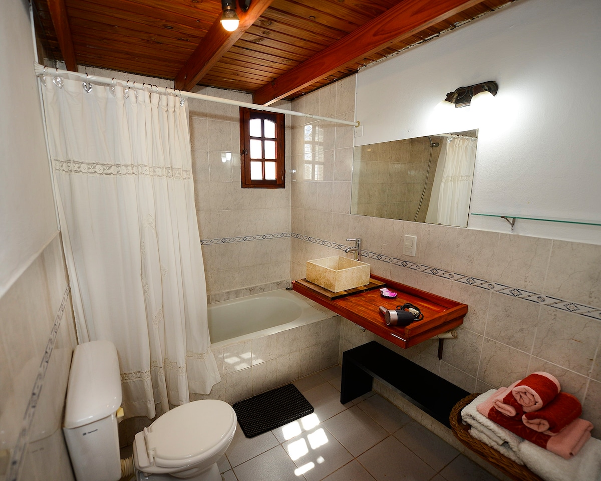 The bathroom features a bathtub and a wooden countertop with a stone sink. Light-colored tiles surround the space, complemented by a mirror above the counter. Towels are neatly arranged on a black bench, with a window providing natural light through decorative grilles.